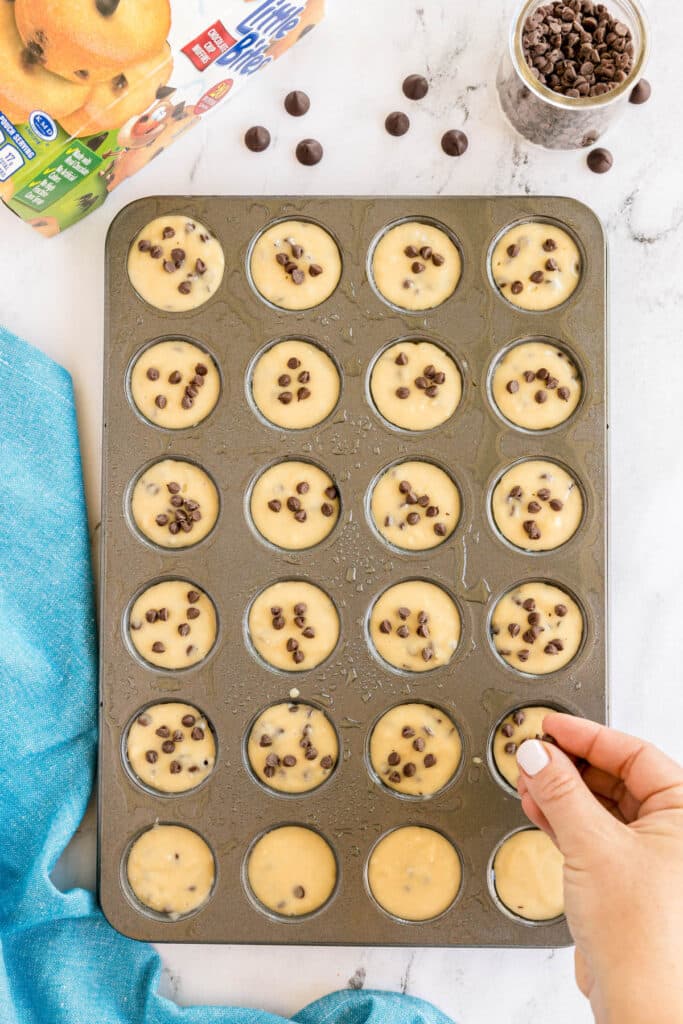 Mini muffin pan filled with chocolate chip batter, some being topped by hand.