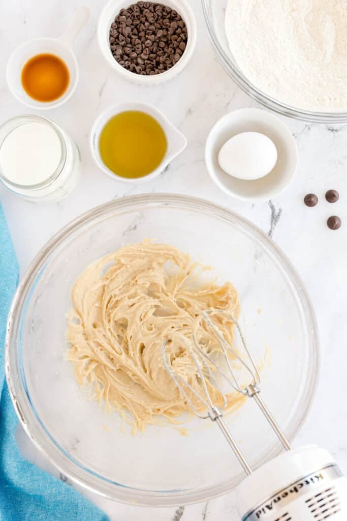 A mixing bowl filled with creamed ingredients, surrounded by bowls of chocolate chips, flour, an egg, oil, milk, and vanilla on a white marble surface.