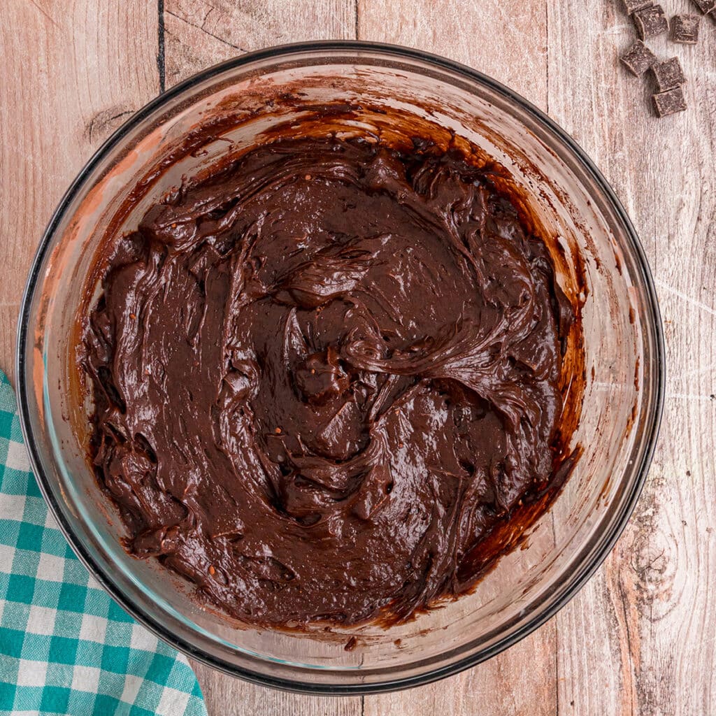 Thick chocolate brownie batter in a glass bowl on a wood surface.
