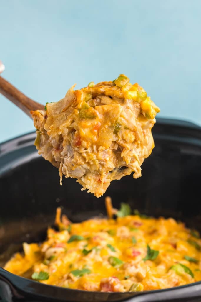 A wooden spoon is lifting a large scoop of creamy, cheesy King Ranch chicken from a black slow cooker.