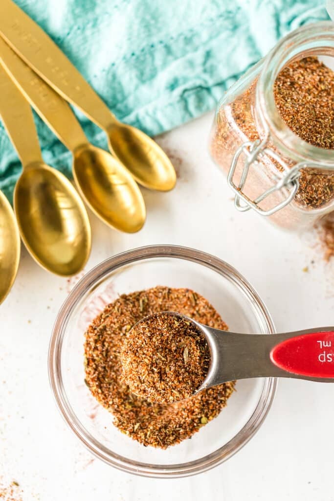 Measuring spoon filled with Cajun seasoning held over a bowl, next to golden spoons.
