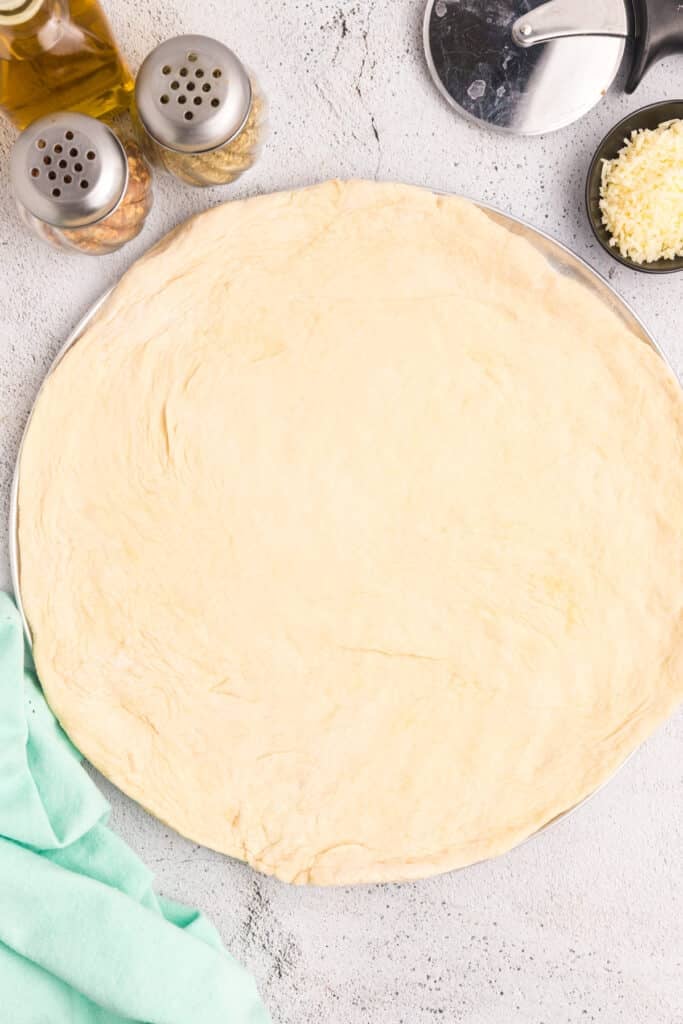 Unbaked pizza dough on a round tray with cheese, a pizza cutter, and condiments nearby.