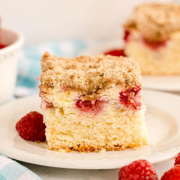 A close-up of a slice of raspberry coffee cake with a golden crumb topping on a white plate.