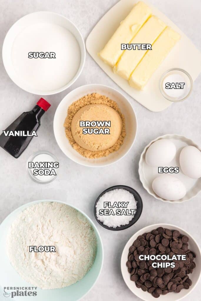 Ingredients for chocolate chip cookies arranged on a white surface, each labeled with text including butter, eggs, flour, and chocolate chips.