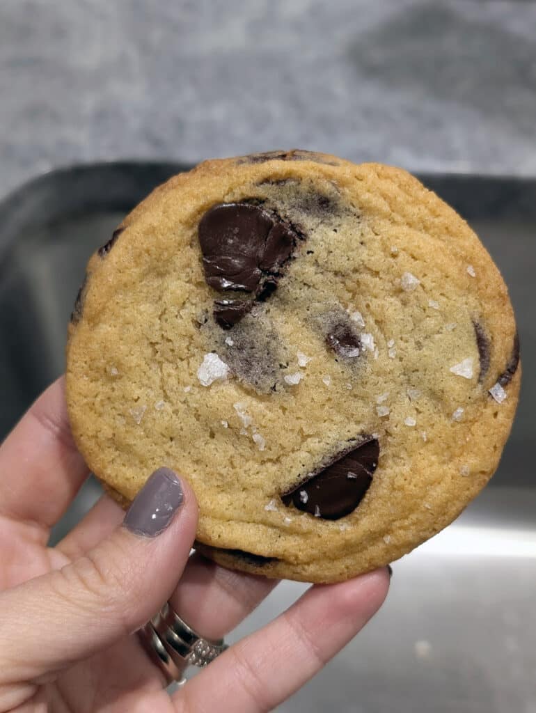 A hand holding a chocolate chip cookie topped with flaky sea salt, with a blurred kitchen sink in the background.