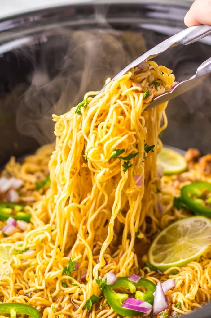 Tongs lifting steaming birria ramen from a slow cooker.