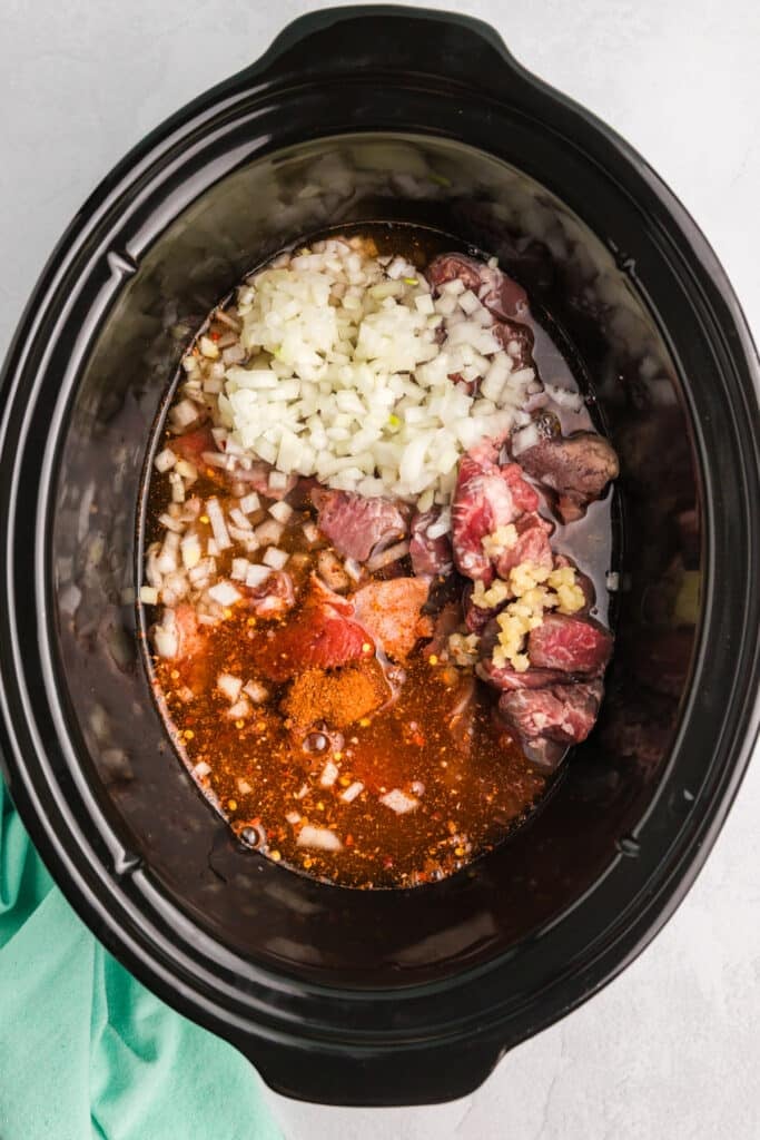 Raw ingredients in a slow cooker including beef, onion, garlic, and spices.