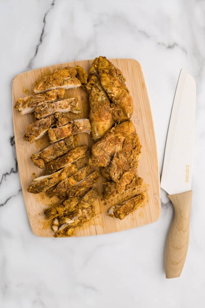 Sliced cooked chicken with seasoning on a cutting board next to a knife.