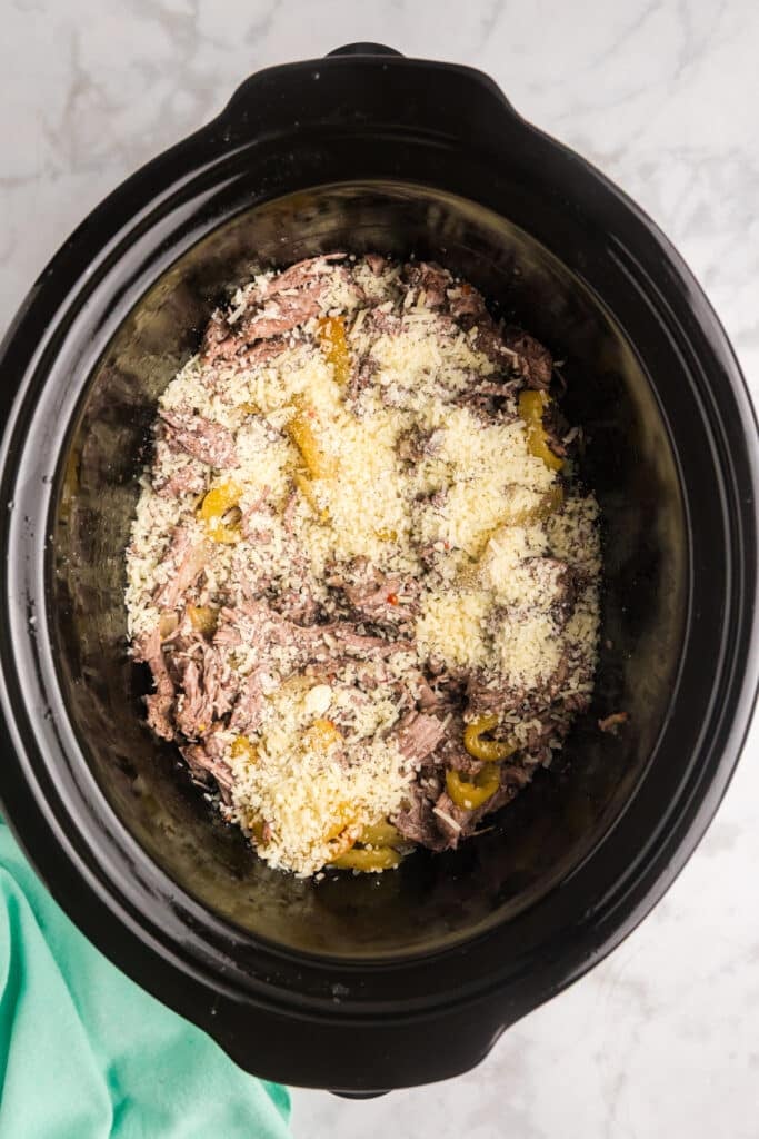 Shredded beef and yellow peppers in a crockpot topped with grated Parmesan cheese.