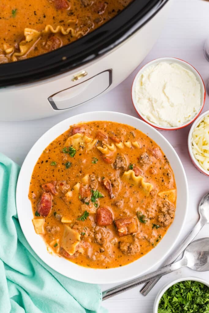 A bowl of lasagna soup topped with parsley sits beside a white slow cooker filled with more soup.