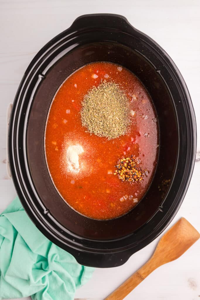 Slow cooker filled with tomato soup base and piles of seasoning.