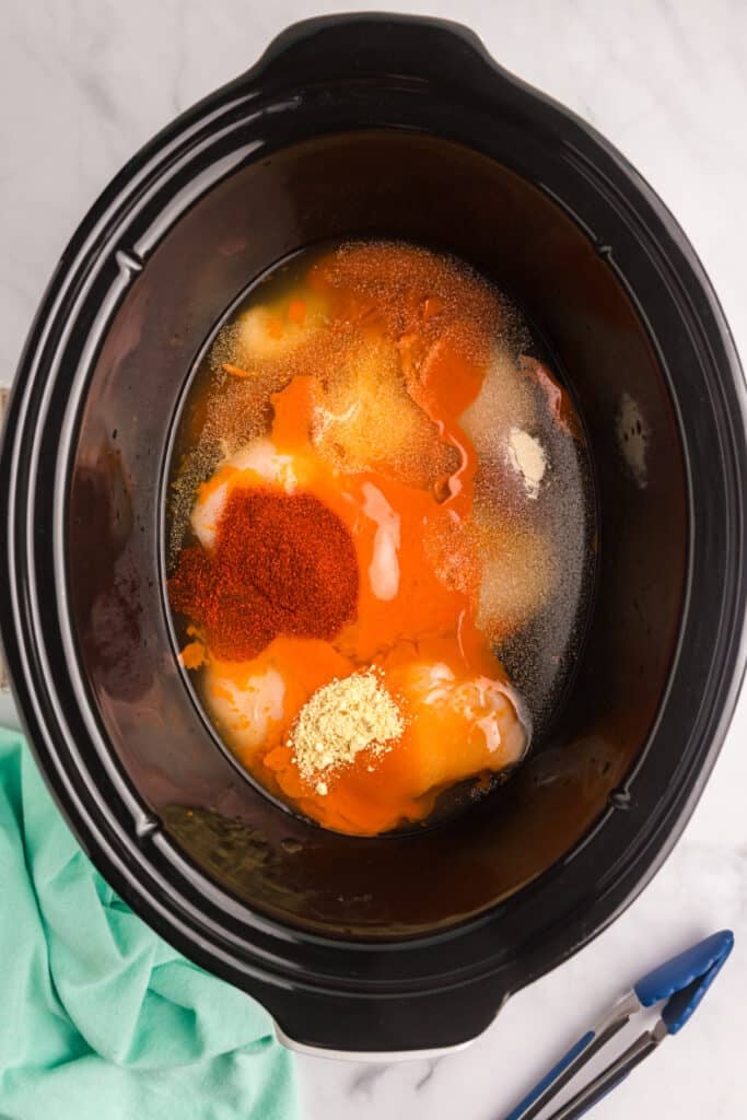 Raw chicken breasts with buffalo sauce and spices in a slow cooker.