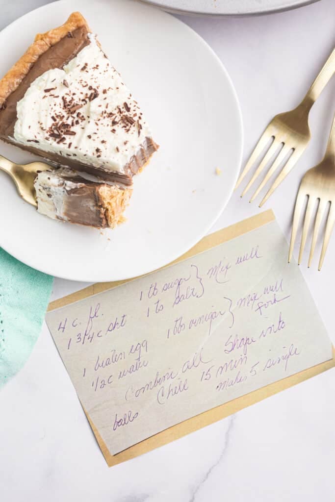 Slice of chocolate cream pie on a white plate next to a handwritten recipe card.