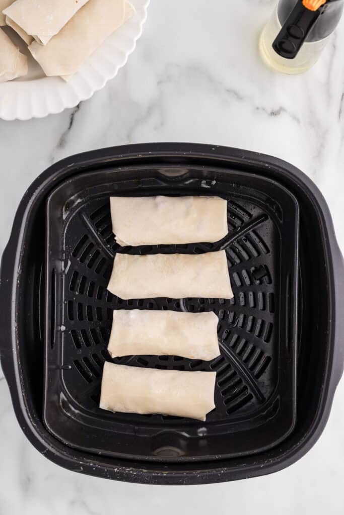 Four uncooked egg rolls placed in an air fryer basket.