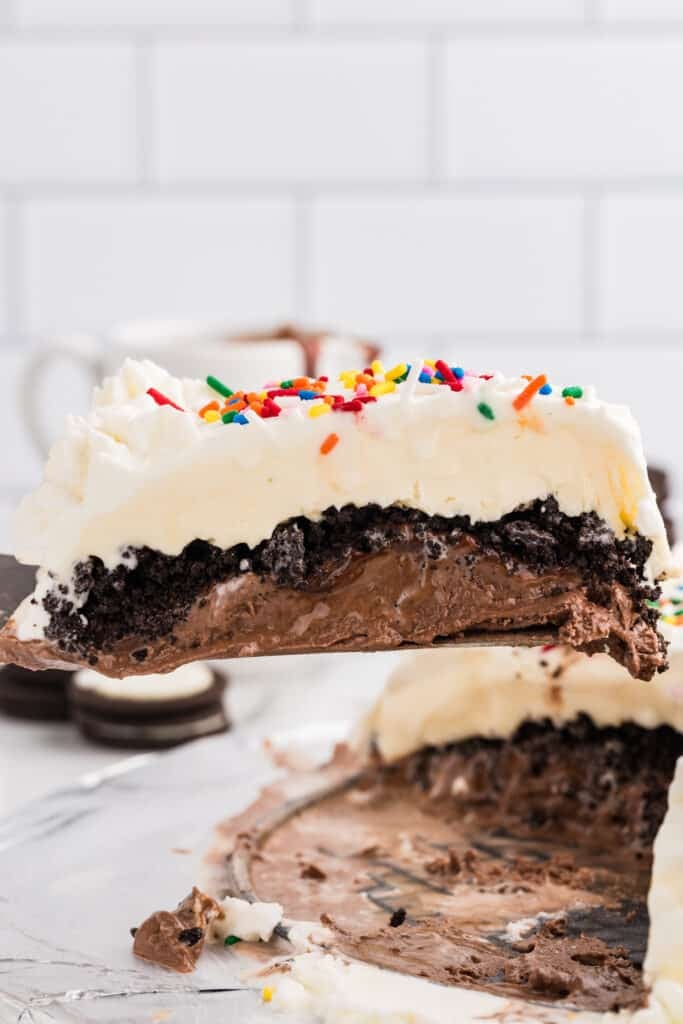 Slice of ice cream cake lifted from the pan, showing layers of vanilla ice cream, cookie crumble, fudge, and chocolate ice cream.