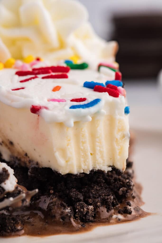 Close-up of a cake slice with white frosting, rainbow sprinkles, cookie crumble, and melting fudge.