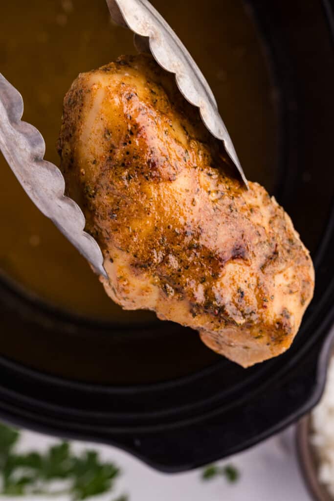 Tongs lifting a seasoned chicken breast from a slow cooker.