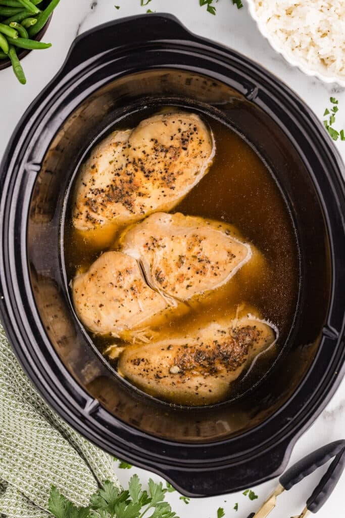 Slow cooker filled with cooked chicken breasts in broth.