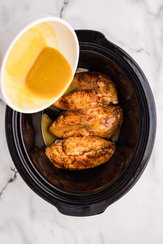Honey mustard sauce being poured over browned chicken in a slow cooker.