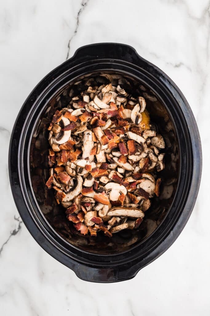 Sliced mushrooms and bacon pieces layered in a slow cooker.