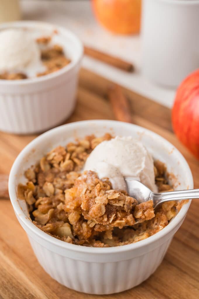 Apple crisp in a white ramekin topped with vanilla ice cream and a spoon inside.