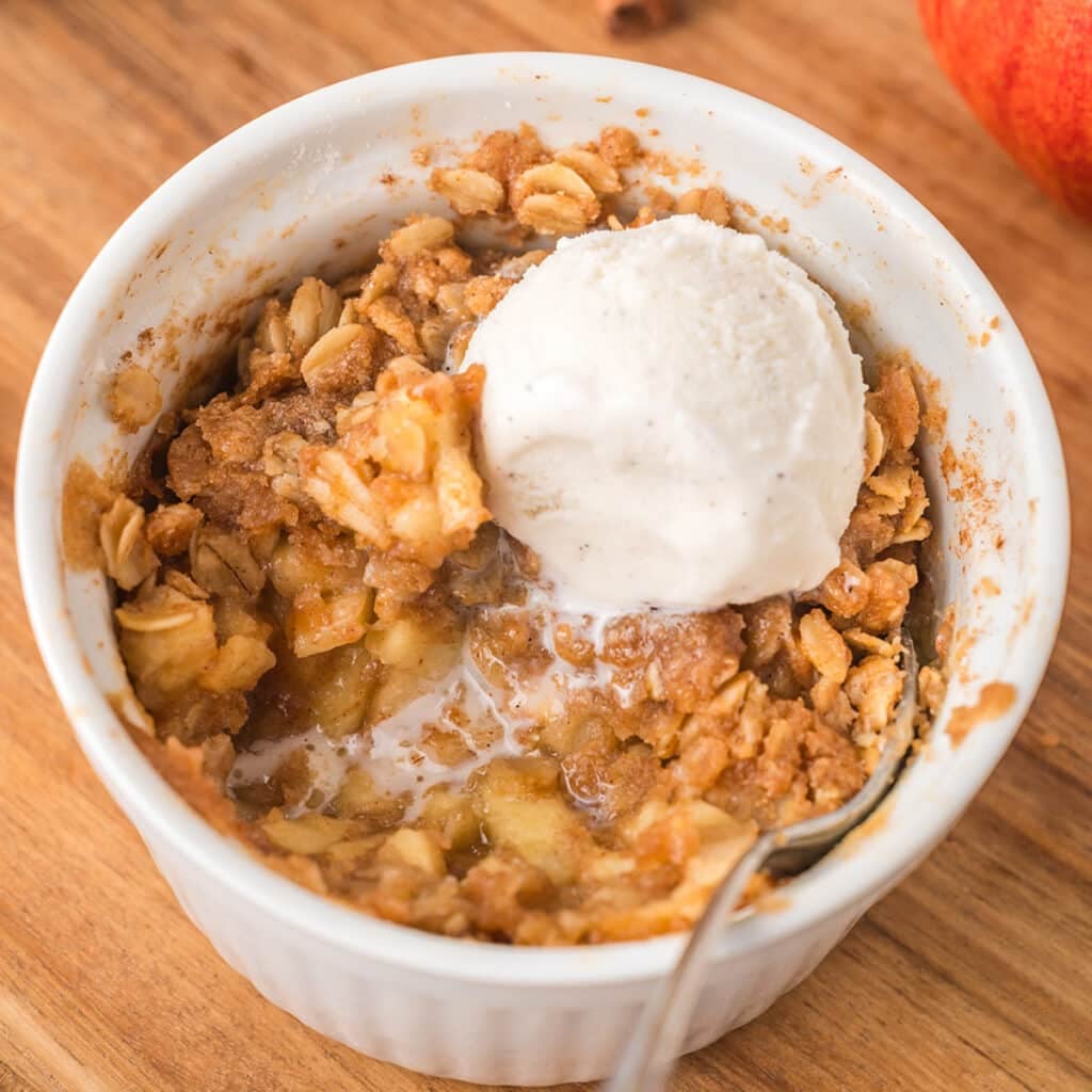 Apple crisp topped with vanilla ice cream in a white ramekin on a wooden board.