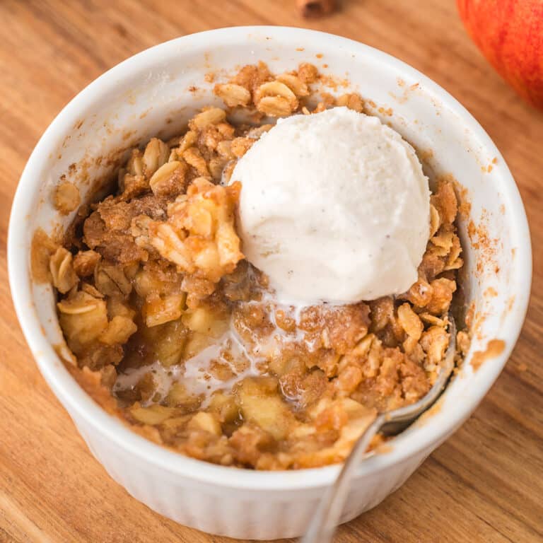 Apple crisp topped with vanilla ice cream in a white ramekin on a wooden board.