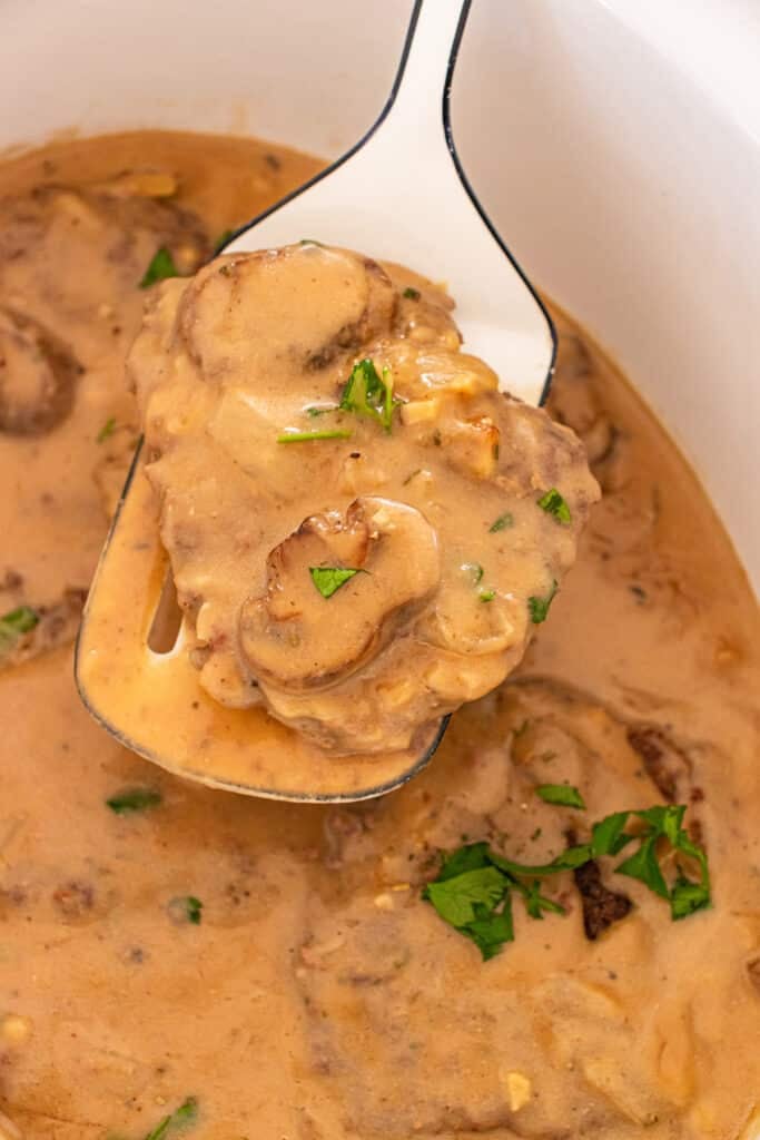 A ladle lifts a piece of cube steak in mushroom gravy from a slow cooker.