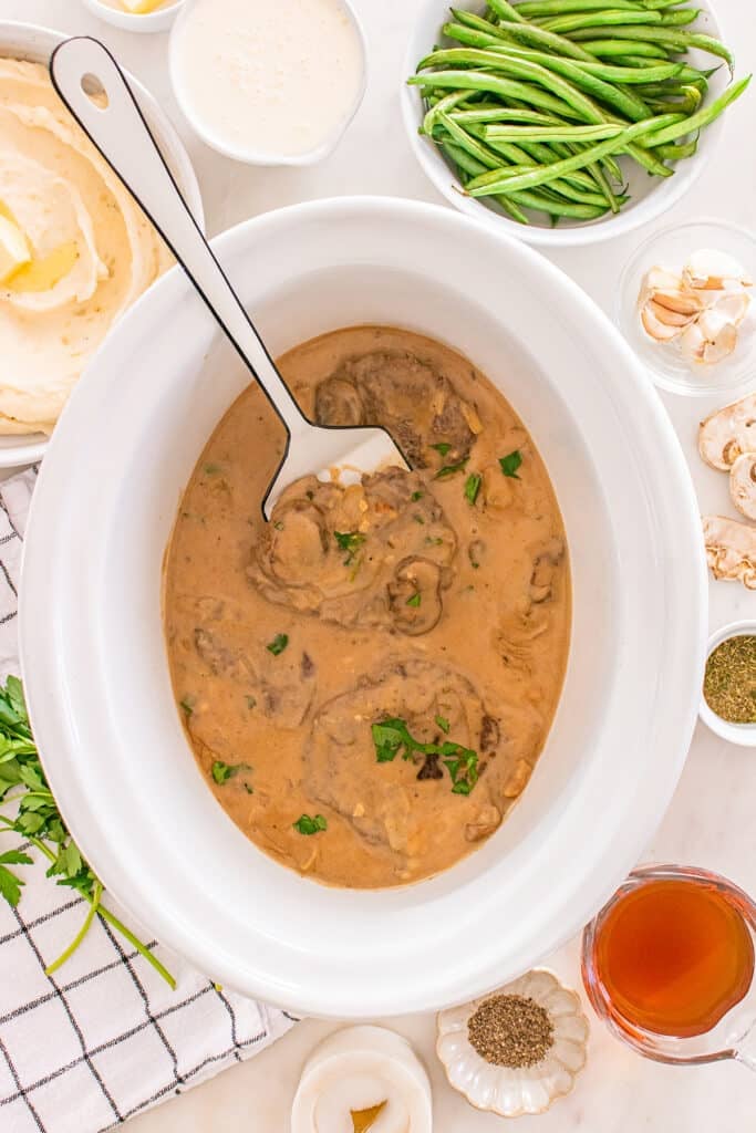 Cube steaks in a creamy brown gravy inside a white slow cooker, topped with parsley.