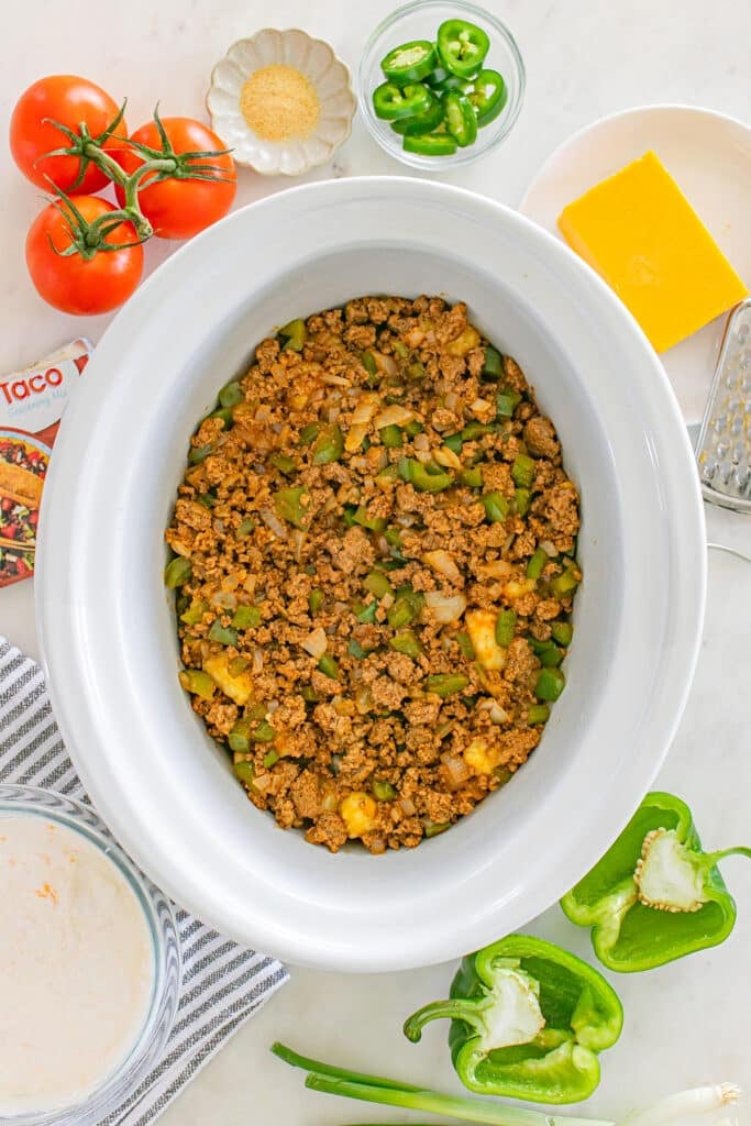 Ground beef and chopped green peppers in a slow cooker surrounded by fresh ingredients.