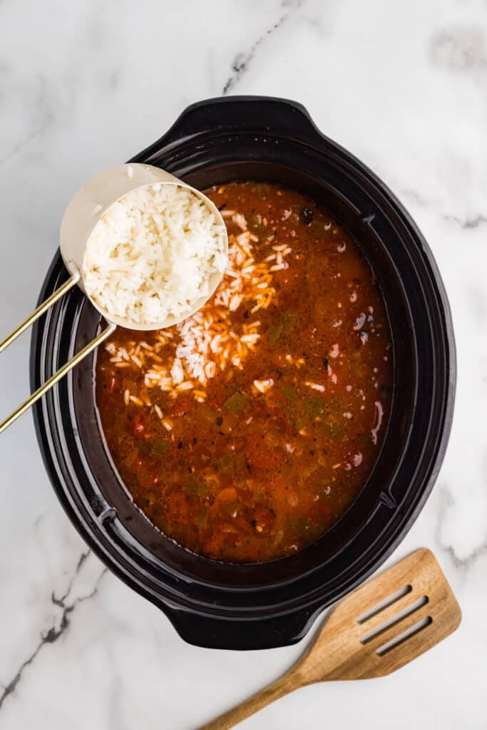 Cooked white rice being poured into a crockpot of tomato-based soup with peppers and beef.
