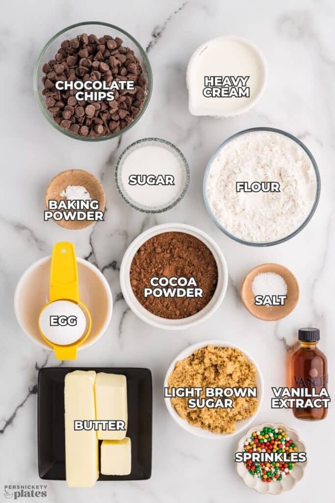 A flatlay of labeled baking ingredients including flour, sugars, cocoa powder, butter, egg, cream, chocolate chips, and sprinkles.
