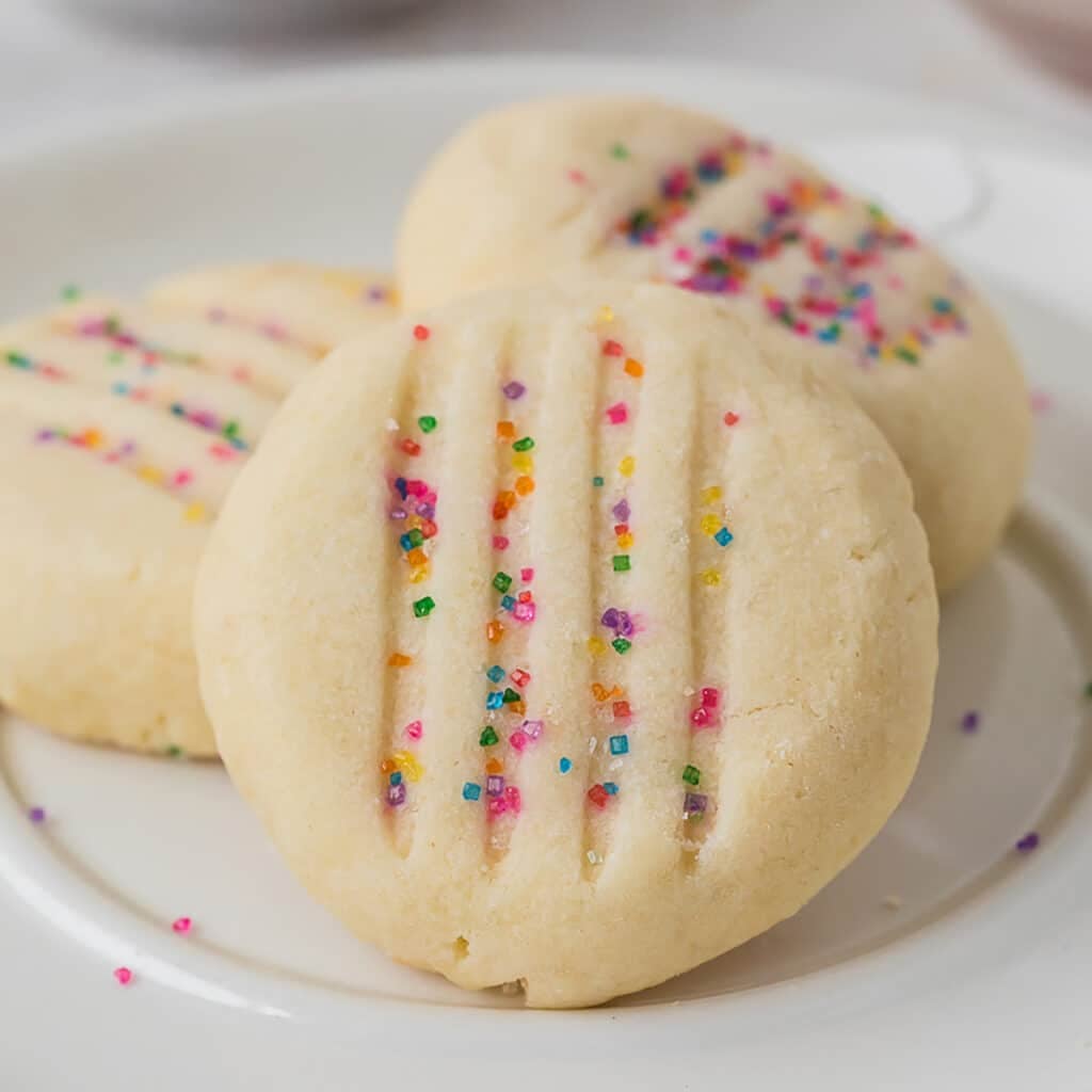 Baked round shortbread cookies with fork marks and rainbow sprinkles on a plate.