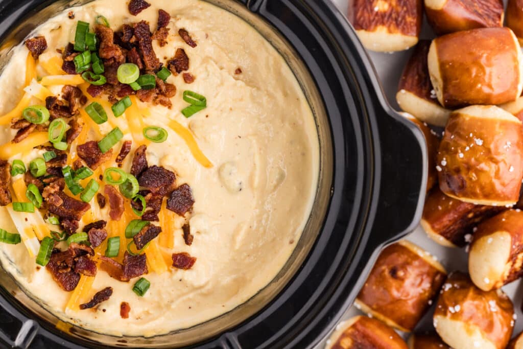Bacon and cheese dip in a slow cooker topped with bacon and green onions beside pretzel bites.