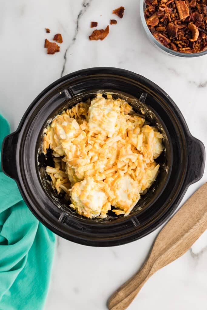 A slow cooker containing large chunks of cream cheese coated with shredded cheese before melting, shown before the ingredients are mixed together.