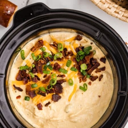 A slow cooker filled with creamy bacon and cheese dip, finished with shredded cheddar cheese, crispy bacon pieces, and sliced green onions on top.