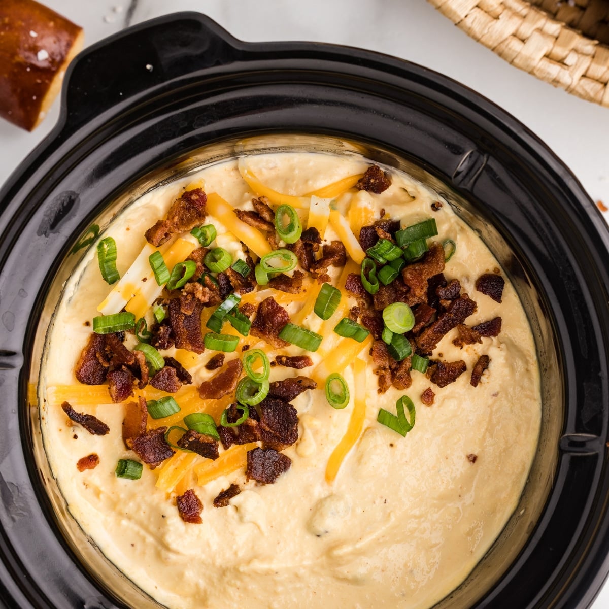 A slow cooker filled with creamy bacon and cheese dip, finished with shredded cheddar cheese, crispy bacon pieces, and sliced green onions on top.