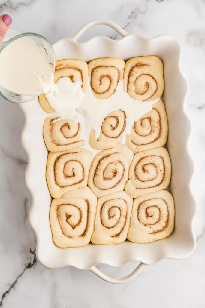 Liquid being poured over unbaked cinnamon rolls in a baking dish.