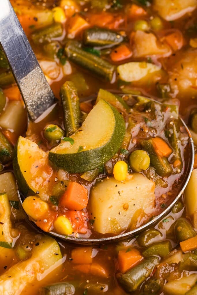 Ladle lifting vegetable soup with potatoes, zucchini, green beans, carrots, and corn.