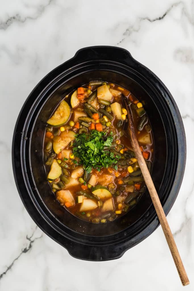 Slow cooker filled with vegetable soup and a wooden spoon.